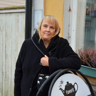 En kvinna står lutad mot en skylt framför en gul husvägg.