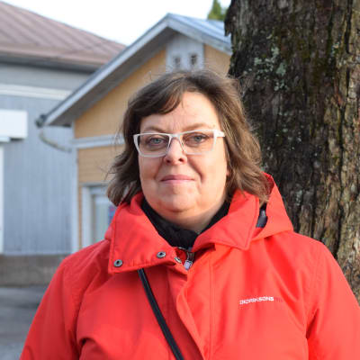 En kvinna med röd jacka och glasögon poserar ute vid ett större träd inne i en stad.