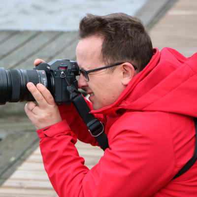 En man står på knä och fotograferar, med en brygga i bakgrunden. 