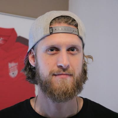 En man med keps på huvudet och långt hår och skägg. Bakom honom syns en röd skjorta upphängd på väggen.