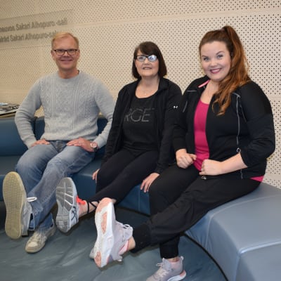 Lääkäri Harri Helajärvi, Taru Lindstedt ja fysioterapeutti Saija Kultala tekevät jumppaa sohvalla.