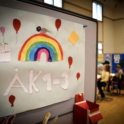 ett klassrum med barn i bakgrunden. I förgrunden en ritad affisch med en regnbåge och texten Åk 1 - 3
