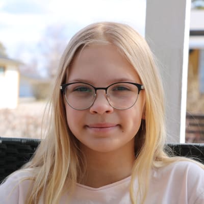 En elvaårig flicka med långt blont hår och glasögon sitter ute på en stol.