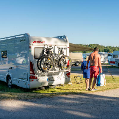 En husbil på en camping. Lättklädda semesterfirare går förbi. 
