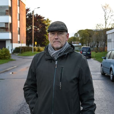 Medelålders man iklädd keps, jacka och halsduk står på gata och tittar in i kameran.