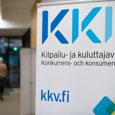 En närbild på en skylt med texten KKV, Konkurrens- och konsumentverket. I bakgrunden syns en människa som går längs KKV:s korridorer.