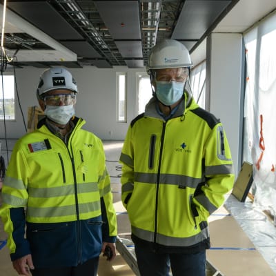 Två män i arbetskläder, hjälm, skyddsglasögon och munskydd står i ett stort rum som är under konstruktion.