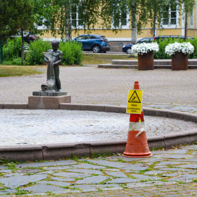 Vattenfontänen i stadsparken i Borgå utan vatten i sig. Bredvid fontänens bassäng står en skylt som varnar för halt botten. 