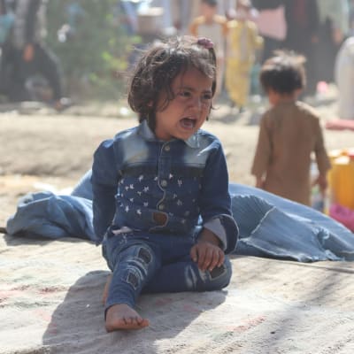 Ett gråtande barn sitter på en matta i ett dammigt flyktingläger