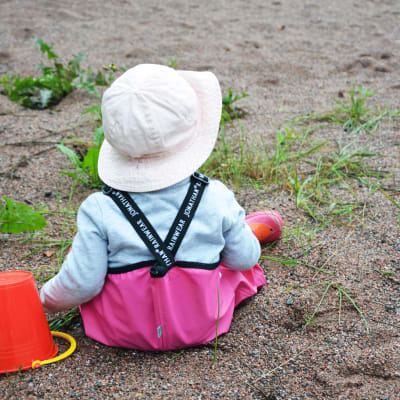 En liten flicka sitter med regnbyxor på i sanden med en hink.