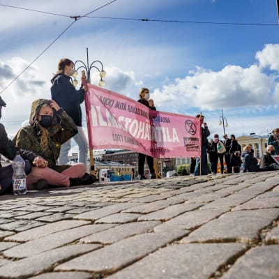 Kaksi Elokapinan mielenosoittajaa istuu kadulla, taustalla banderolli jossa lukee ilmastohätätila..