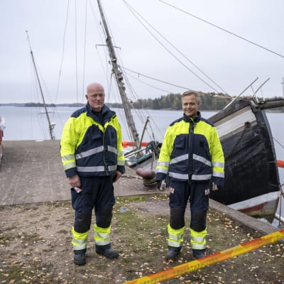 Jörgen Engroos och Fredrik Kevin står på kajen bredvid en stor trålare som ligger halvvägs under vattenytan.