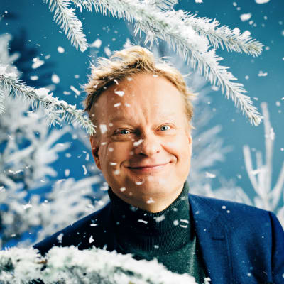 Pianisti Iiro Rantala katsoo puolilähikuvassa hymyillen kameraan, ympärillään luminen kuusi ja sen "hiutaleita".