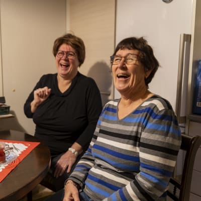 Två kvinnor sitter vid ett köksbord och skrattar hjärtligt. Det är dukat med julduk, glögg och adventsljusstake i fönstret.