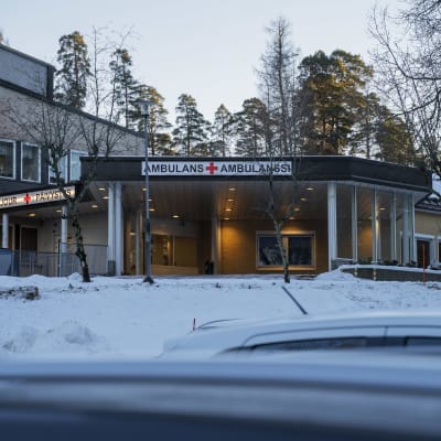 En sjukhusbyggnad med en stor skylt där det står ambulans på svenska och finska. I förgrunden syns taket av några bilar.