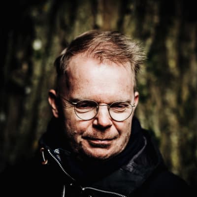 En man i runda glasögon och svart jacka står framför en trädstam.