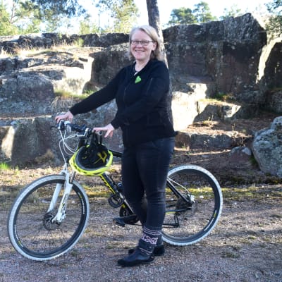 Blond kvinna står med sportig cykel vid ett berg. Hon är klädd i svart, på cykeln hänger en gul cykelhjälm.