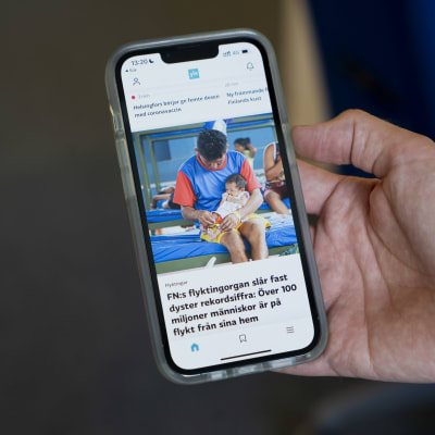 En hand håller upp en mobiltelefon med den svenskspråkiga startvyn för Yle-appen. Skärmen visar en nyhet om att över 100 miljoner människor i världen är på flykt.