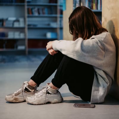 En flicka sitter lutad mot en bokhylla på golvet.