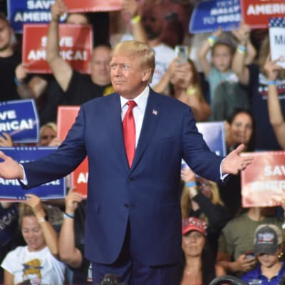 På bilden syns ex-presidenten Donald Trump på ett av sina politiska evenemang. I bakgrunden står hans anhängare med skyltar i blått, rött och vitt där det står "Save America". 