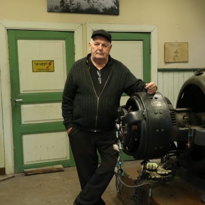 En man med keps på huvudet står och lutar sig mot en gammal generator.