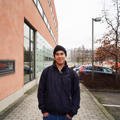 En ung man i jacka och mössa ler in i kameran framför parkerade bilar och en husvägg.