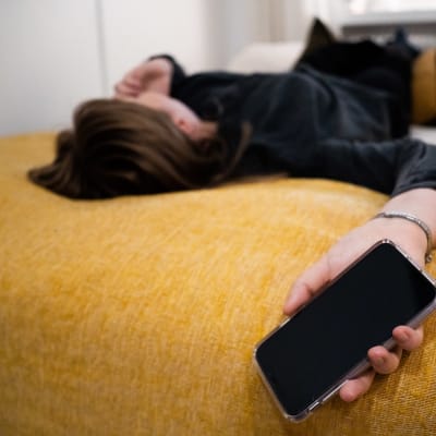 En person med långt hår ligger på en säng och håller i en telefon.