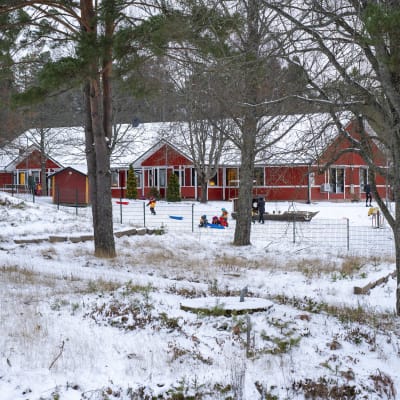 Ett rött hus skymtar bakom träd. Det är snö på gården och någrab arn åker pulka. Det är Österbo daghem.
