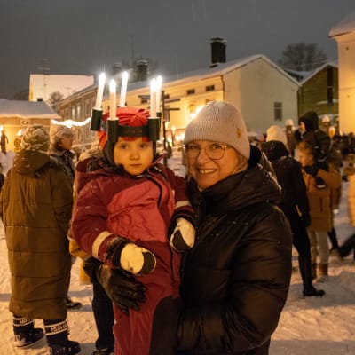 En liten flicka med luciakrona i famnen på en kvinna. Båda ler och tittar in i kameran. 