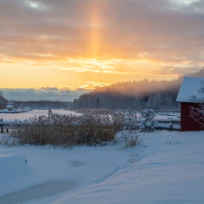 Gammal röd träbyggnad vid kanten av en å i ett vintrigt landskap. 