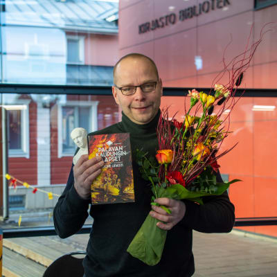 En glad man håller i en blombukett, en bok och en liten vit Runebergsstaty.