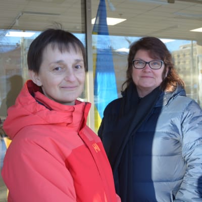 Två kvinnor står utomhus och tittar in i kameran, mellan dem skymtar en ukrainsk flagga.