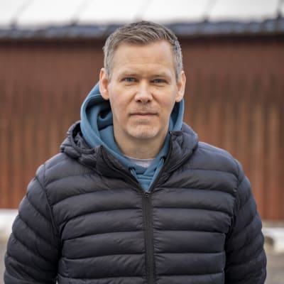 En man med gråsprängt kort hår och mörkblå tunn dunjacka står ute. I bakgrunden en byggnad med rödmålad träfasad.