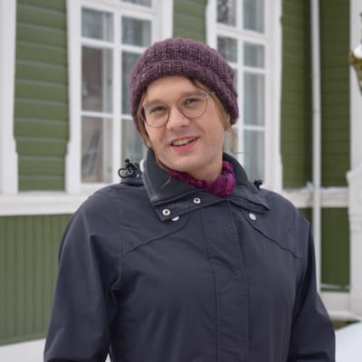 Petra Lönnberg står framför en grön husfasad iklädd mössa och jacka. Trädet i bakgrunden är delvis snötäckt. 