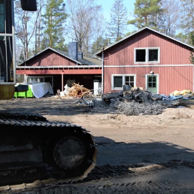 Grävmaskin i förgrunden och en stor röd träbyggnad i rivningsskede i bakgrunden.
