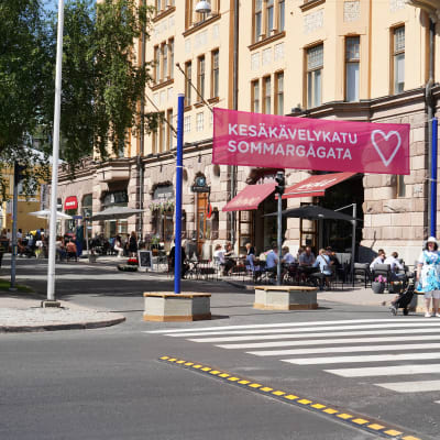 Ett stort övergångsställe framför en skylt där det står sommargågata. På gågatan sitter människor på uteserveringar.