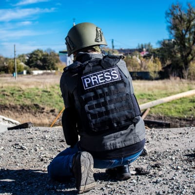 En fotograf iklädd skyddshjälm och skyddsväst där det står "press" på ryggen hukar för att ta en bild i Bachmut.