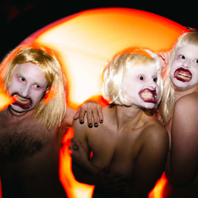 Tre nakna skådespelare med uppspärrade munnar och blonda peruker. 