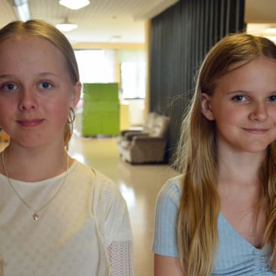 Två flickor i 11-årsåldern står i en skolkorridor och tittar in i kameran.