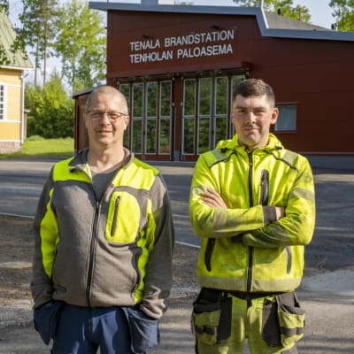 Två arbetsklädda (gula och gråsvarta kläder) män framför en ny brandstation. Tenala brandstation.
