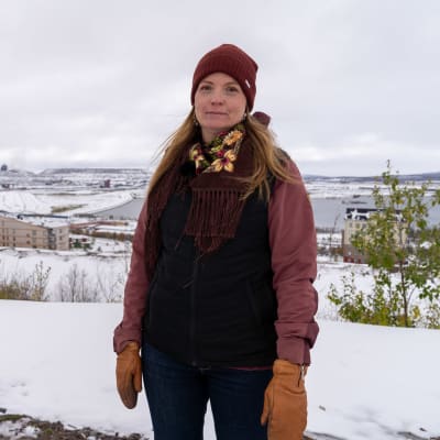 Karin Kvarfordt Niia står med en röd mössa på och halsduk och tittar mot kameran. I bakgrunden hus och ett berg.