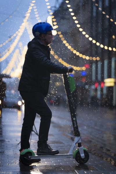 En elsparkcykelåkare med blå hjälm står i skymningen i regn vid Alexandersgatans julbelysning i Helsingfors.