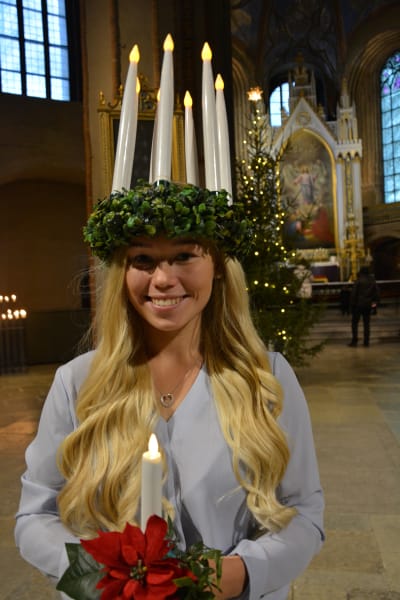 Åbos lucia 2020 och 2021, Janina Karrento, en dam med långt ljust hår och en ljuskrona på huvudet, ler mot kameran. Hon står i Åbo Domkyrka med en stor julgran i bakgrunden.