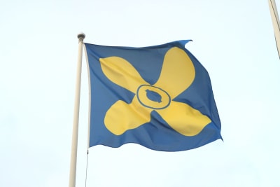 Kimitoöns blåa flagga med en gul propeller i mitten mot en ljusblå himmel.