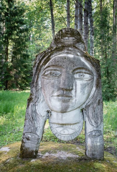 Harmaa patsas, jossa kasvot, taustalla koivikkoa