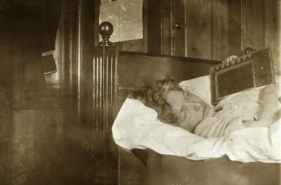 Edith Södergran ligger på sjukbädd med typ en radio ? på bröstet. Ser in i kameran. Kudde under huvudet.