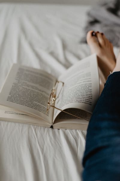 Någon läser en bok i sängen med glasögonen liggande i boken.