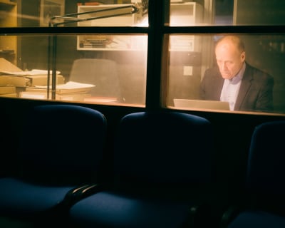 Iäkäs mies työskentelee lasiseinän takana pöytälampun valossa. Lasiseinän edustalla näkyvät arkistohuoneen tuoleja.