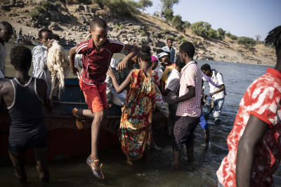 Etiopiska flyktingar korsar en flod på väg till Sudan, i förgrunden en pojke i orange shorts och en röd t-shirt.