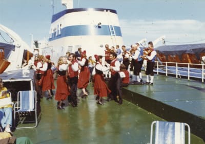 Folkdansare i röda och svarta folkdräkter dansar på däck framför den blåvita skorstenen på en färja som är på väg till Örnsköldsvik.